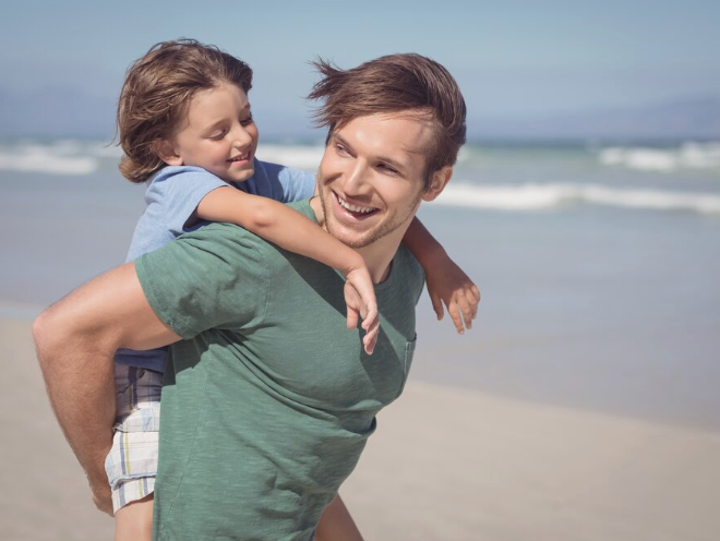 Man walking on beach carrying child on his back.